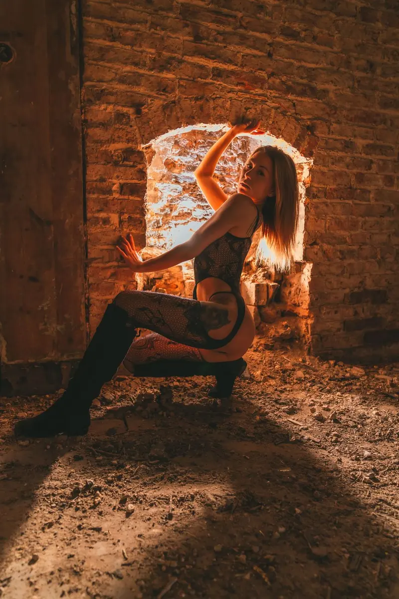 A woman in a black lace bodysuit and black thigh-high boots crouching in a brick room, one hand on the wall, with warm light from a window.