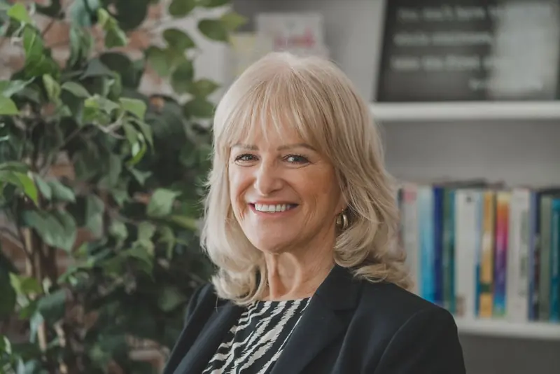 A smiling woman with shoulder-length blonde hair, a black blazer, and a zebra-print top, with a leafy plant and colourful books behind her.