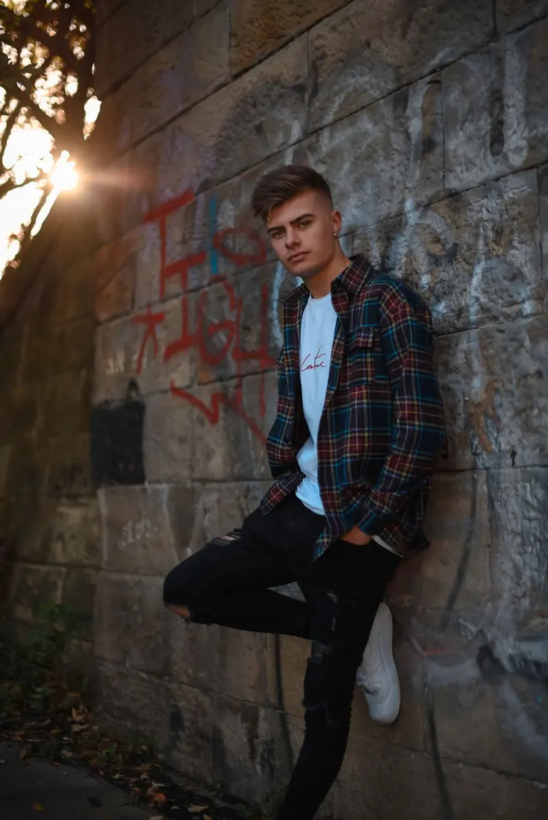 A young man leaning on a stone wall in a blue, red, and yellow plaid shirt, ripped black jeans, and white trainers, with red graffiti in the sunlight.