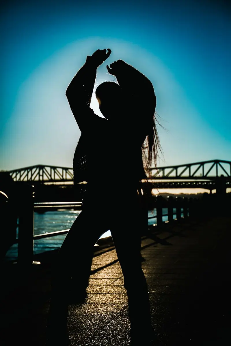A silhouette with both arms raised on a waterfront path, a metal bridge and water behind, under a blue sky.