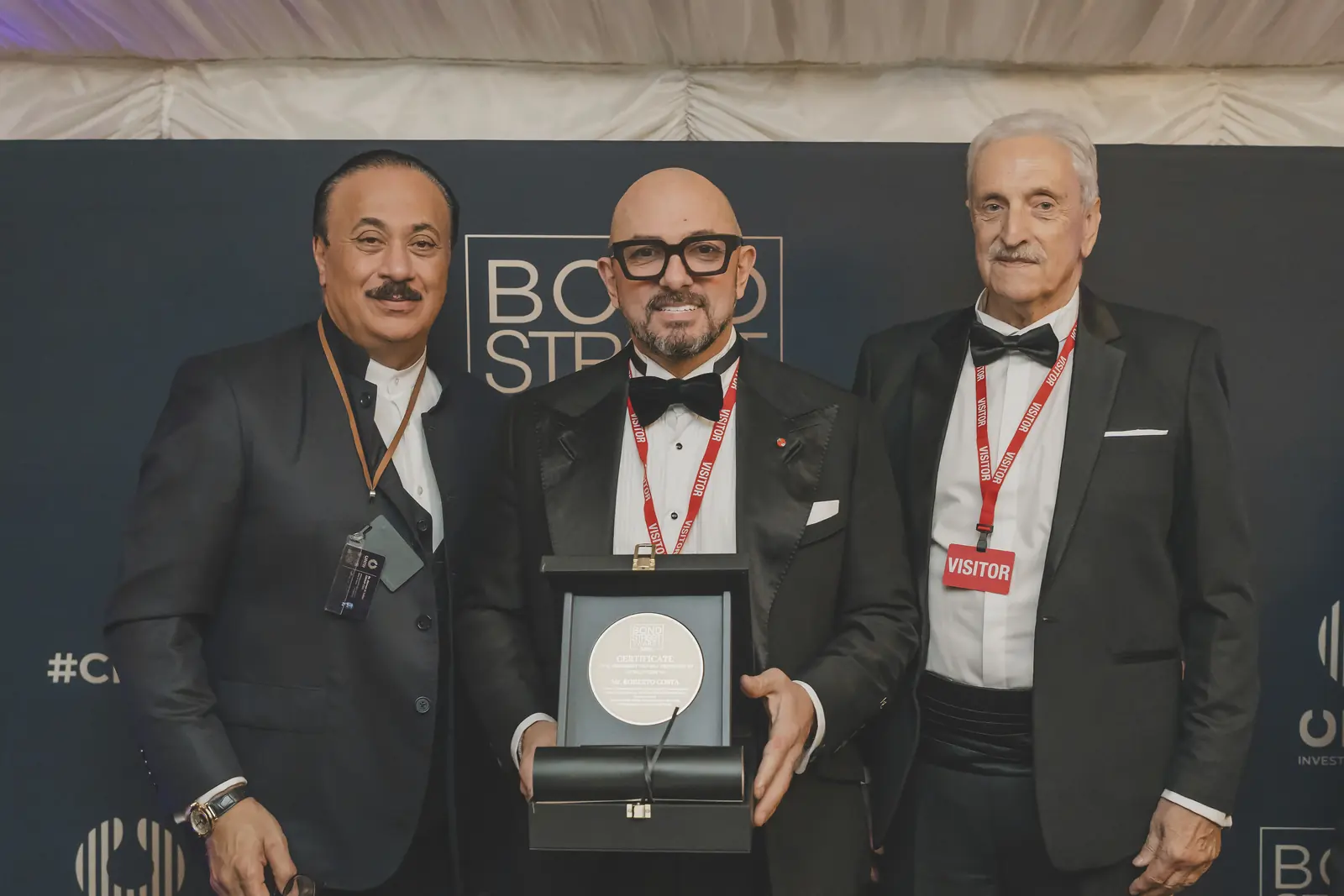 Three men in dark suits at the CNIW/ZAI BSA 2024 awards. The man in the tuxedo holds an open case with a round certificate.