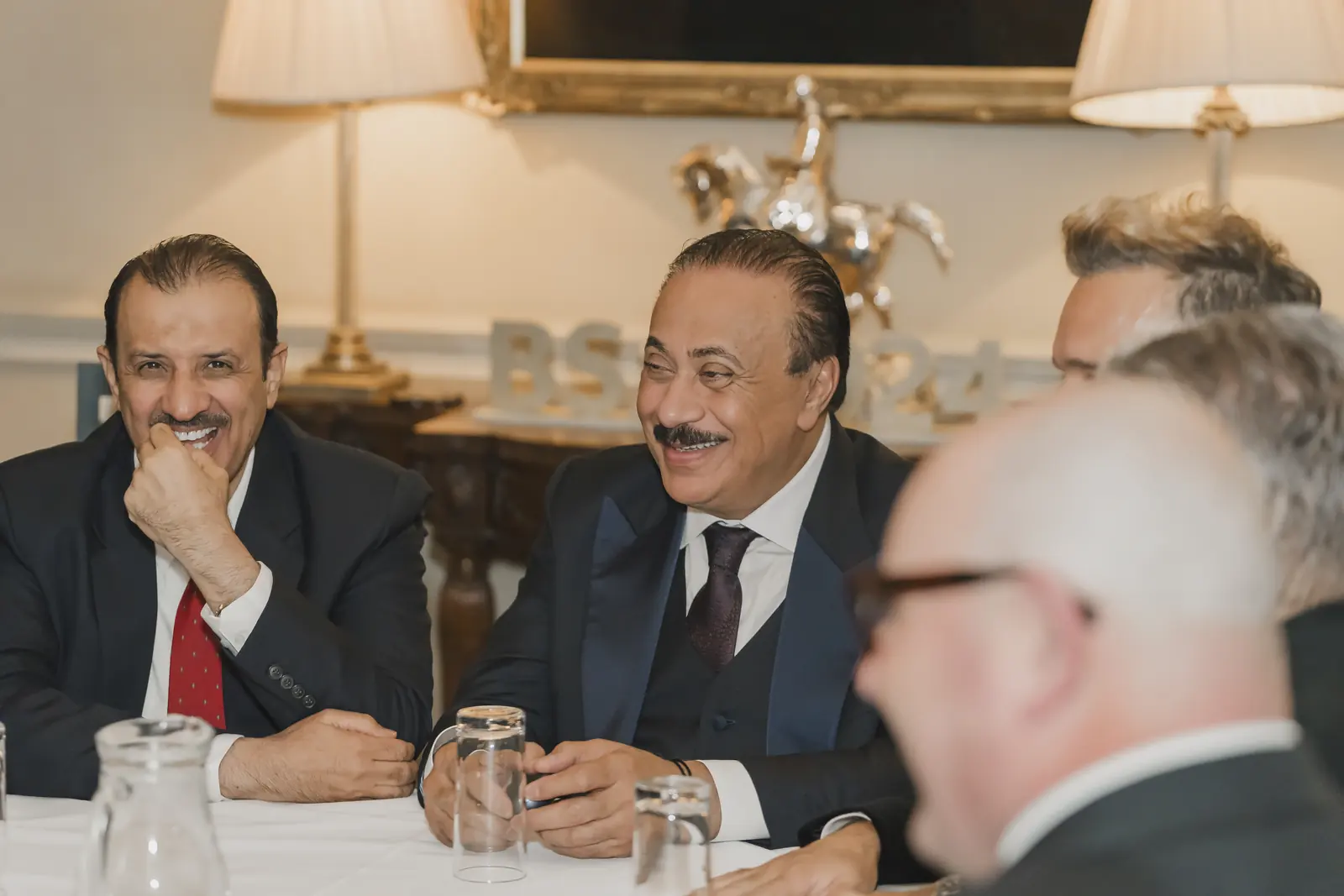 Men in dark suits seated at a white-clothed dinner table at the CNIW/ZAI BSA 2024 awards, with glasses, a water jug, and a silver horse ornament.