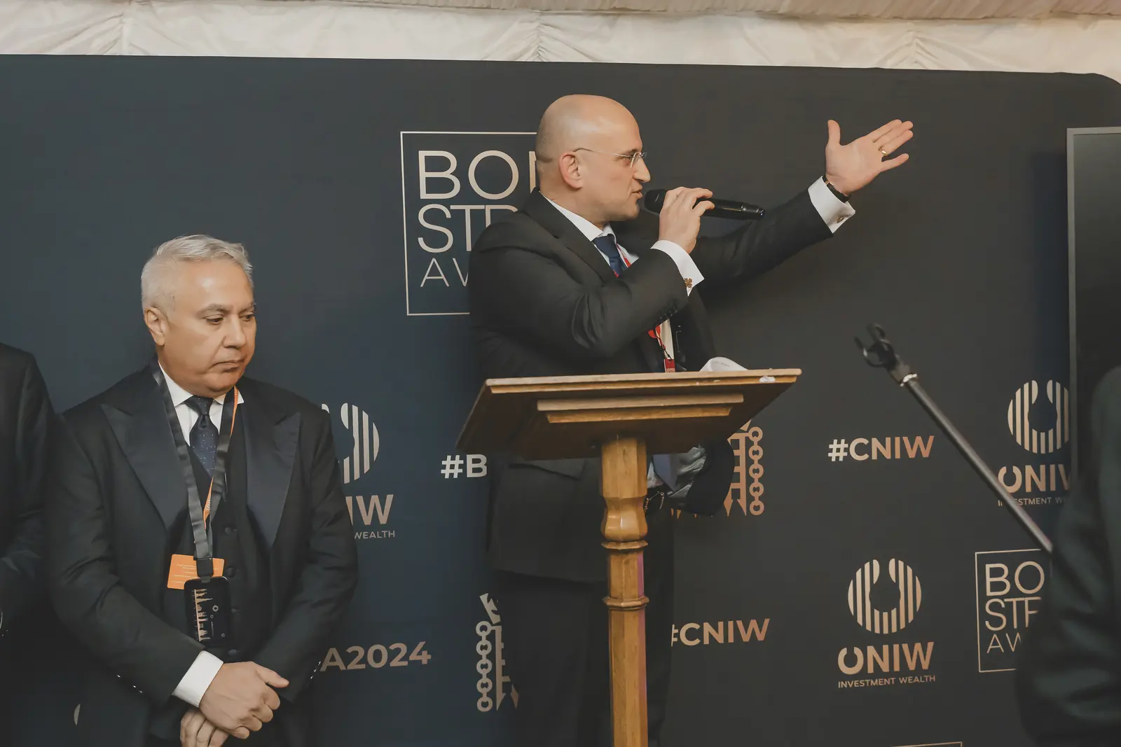 A bald man in a dark suit speaking at a wooden lectern at the CNIW/ZAI BSA 2024 awards, a grey-haired man beside him with his hands clasped.
