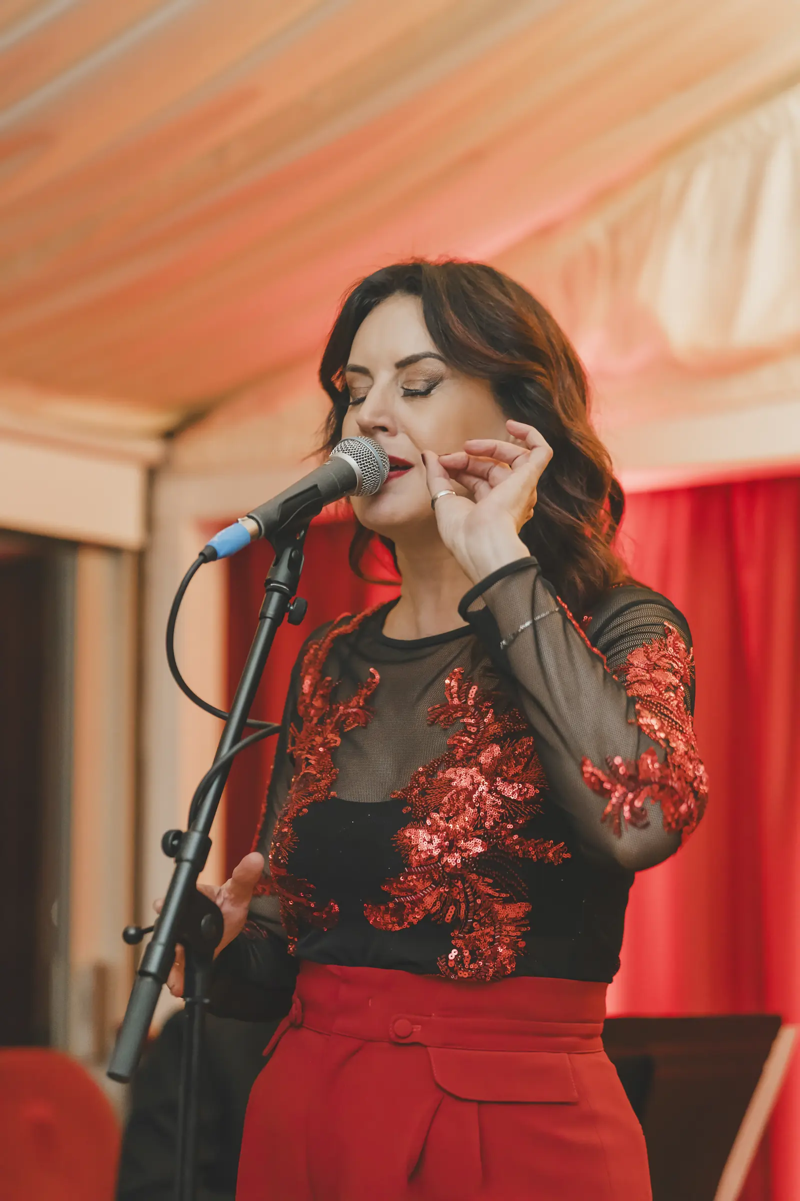 A woman in a black top with red sequins and red trousers singing at the CNIW/ZAI BSA 2024 awards, red curtains behind her.