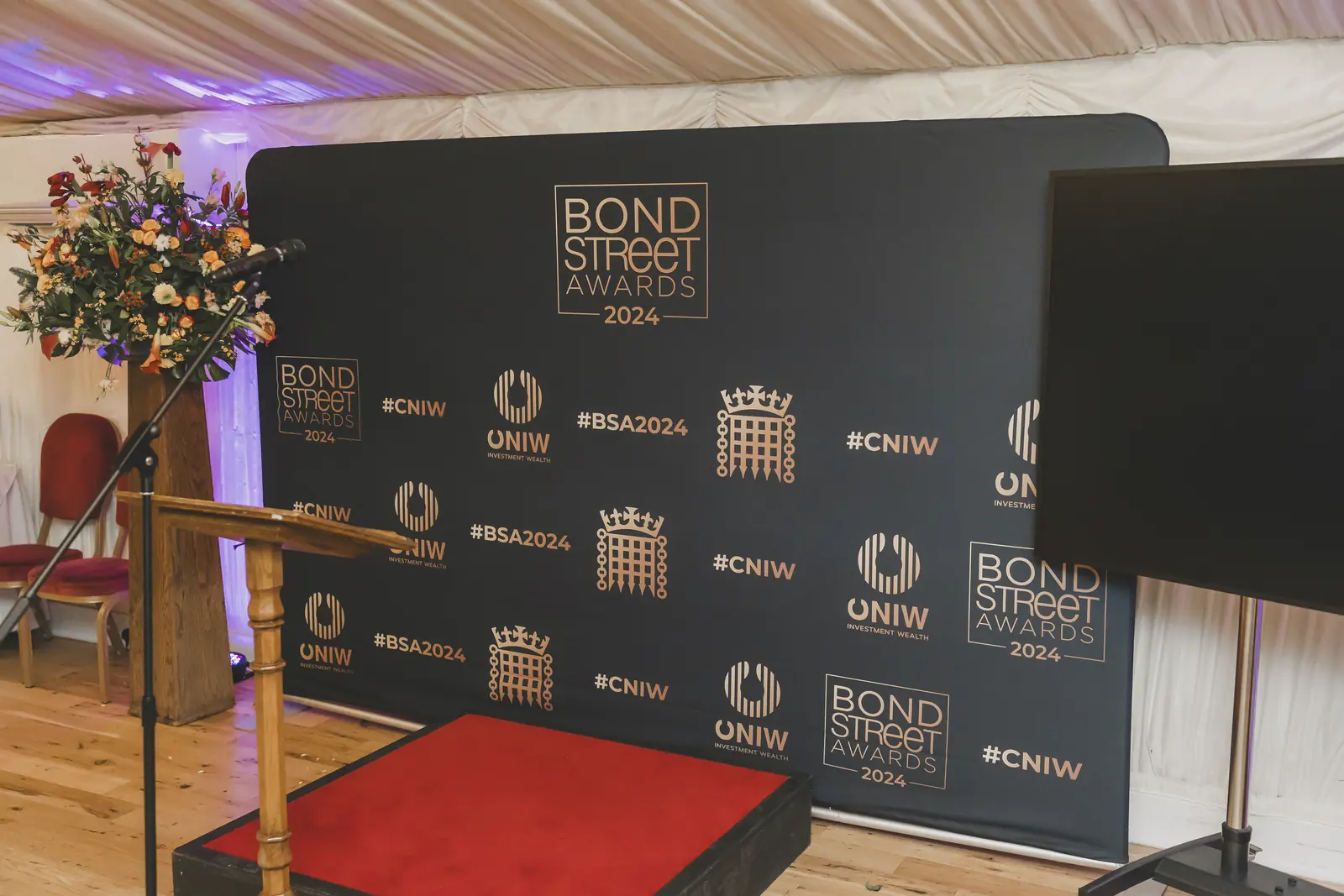 An empty stage under a white tent at the CNIW/ZAI BSA 2024 awards: a black step-and-repeat with gold logos, a wooden lectern, and a red floor. No people.