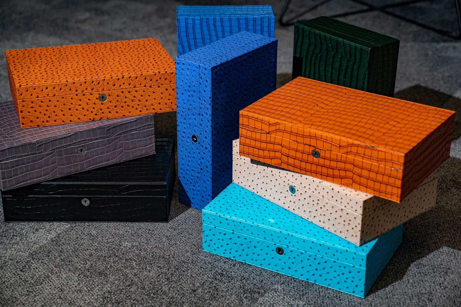 Stacked rectangular boxes in orange, blue, green, grey, black, cream, and turquoise, with metal clasps, on grey carpet. Etier.