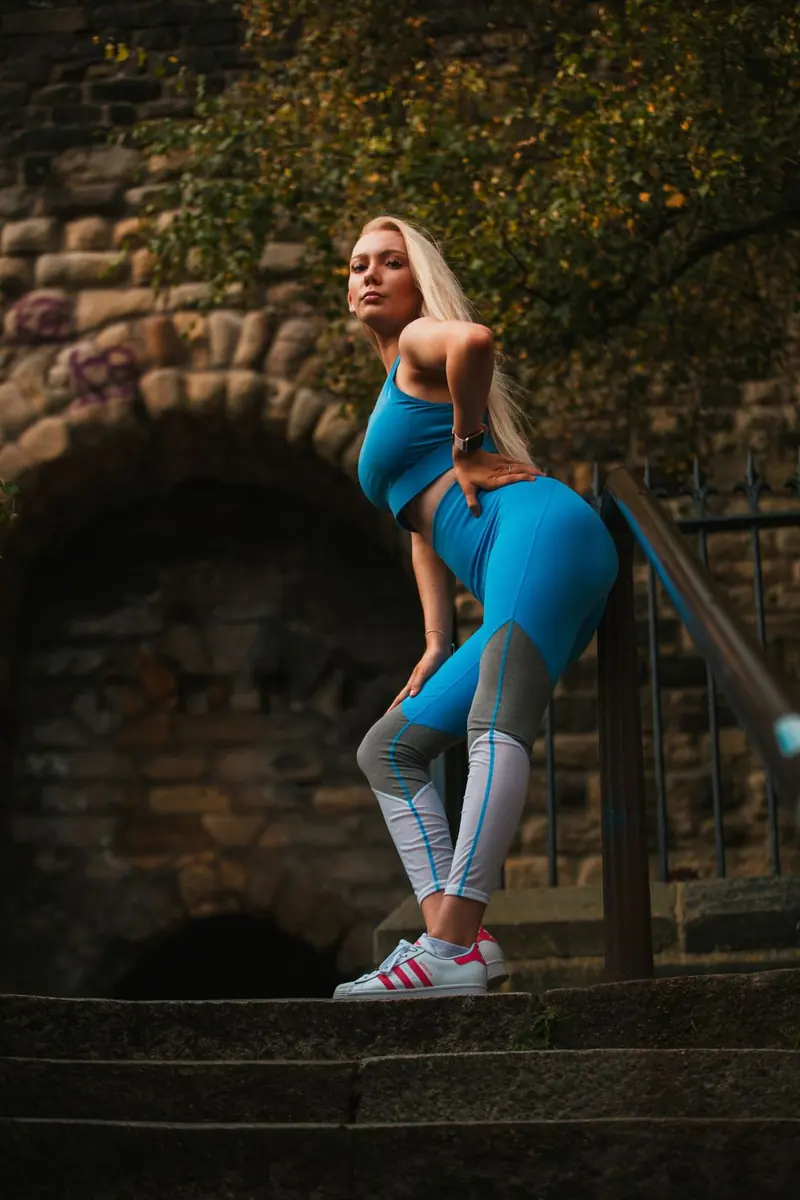 A woman with long blonde hair in a bright blue crop top and blue, grey, and white leggings, on stone steps by a metal rail.
