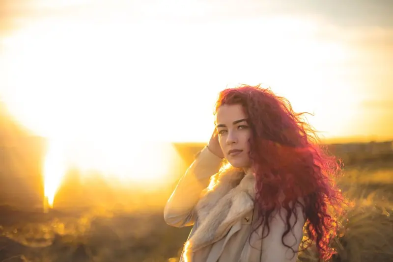 A woman with long curly red hair and a pale coat with a light fur collar, one hand to her head, in a field under a golden sunset.
