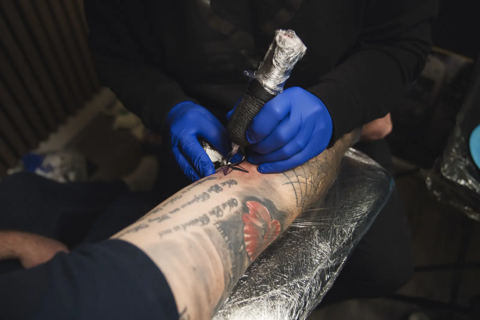 A tattoo in progress at Gym22: blue gloves and a plastic-wrapped machine on a forearm with script and a red flower, over clear plastic.