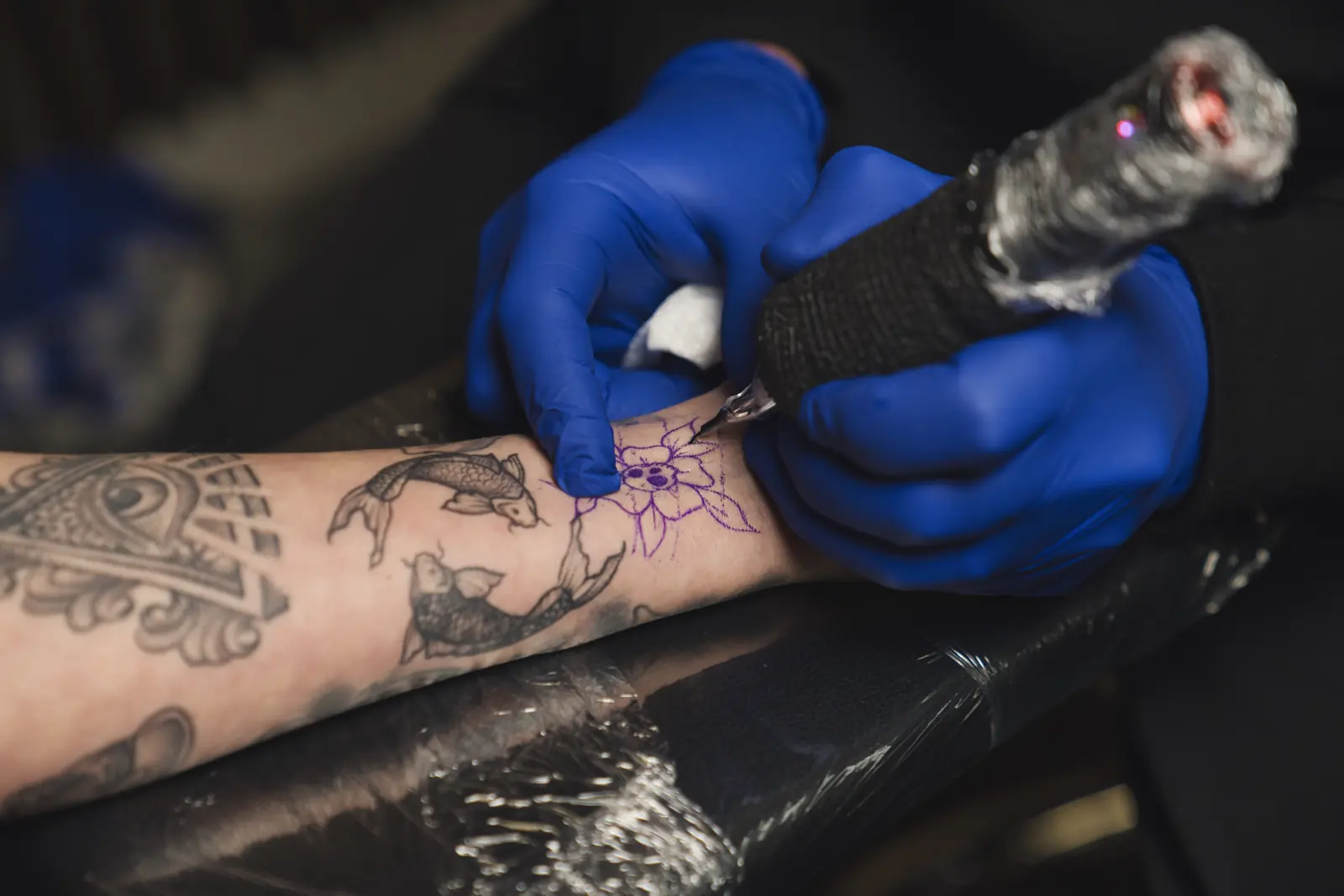 Blue-gloved hands tattooing a purple flower stencil on a forearm that already has black tattoos of fish, an eye, and ornamental patterns. Gym22.