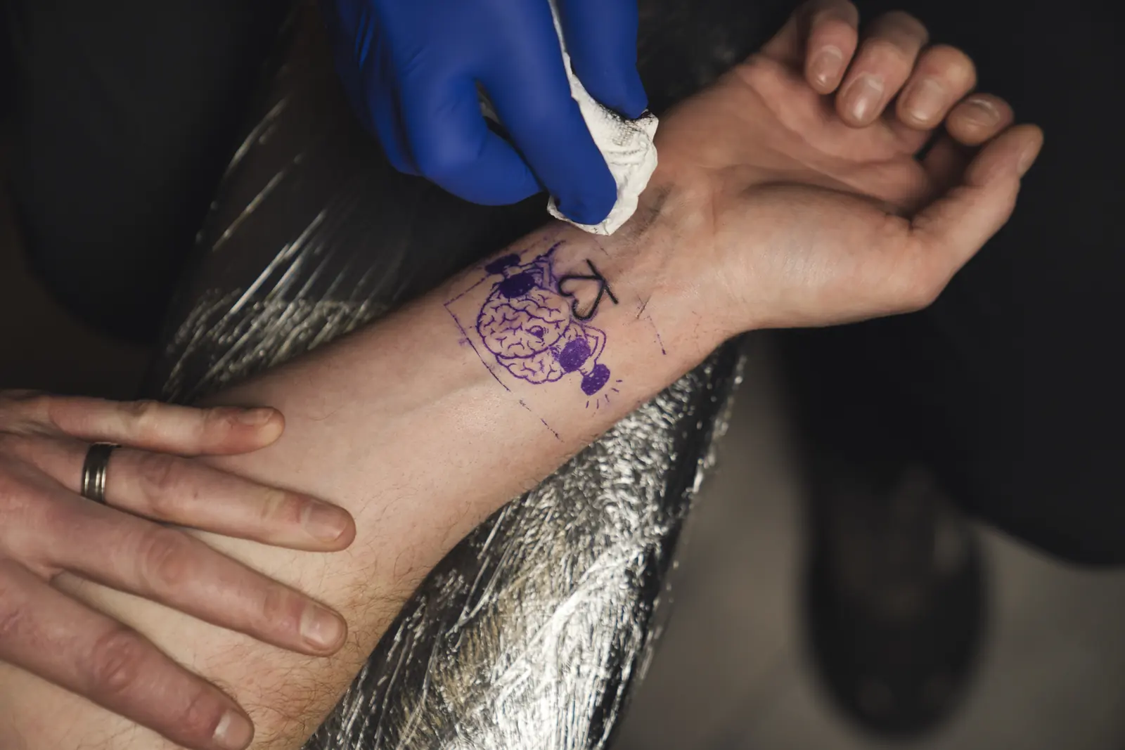 Blue-gloved hands and a white cloth on a forearm at Gym22. The purple stencil is a brain inside a square, with a lightbulb and a heart.