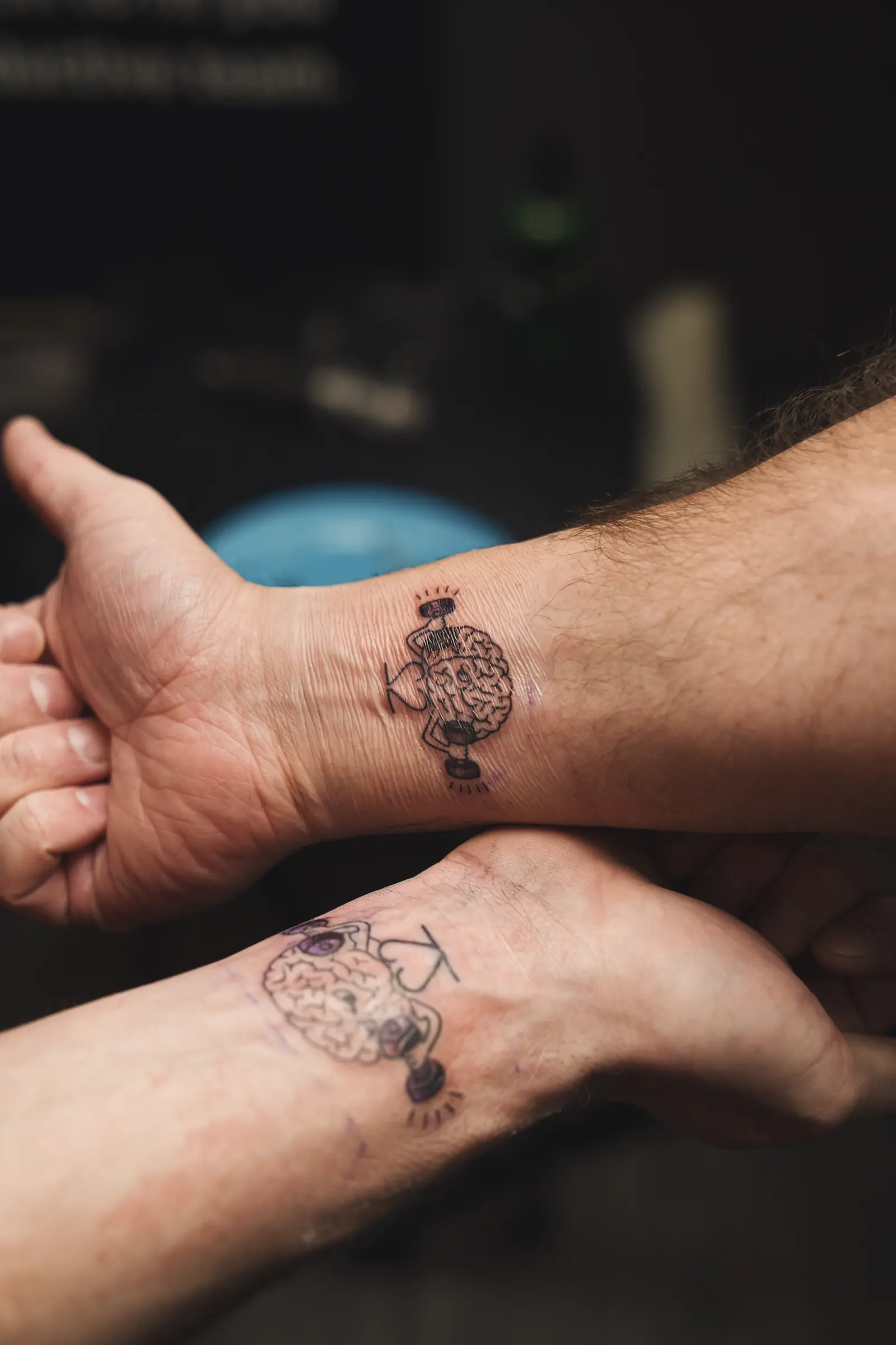 Two inner wrists at Gym22. One tattoo is a cartoon brain lifting dumbbells. The other is a brain lifting weights beside a heart, with purple shading.