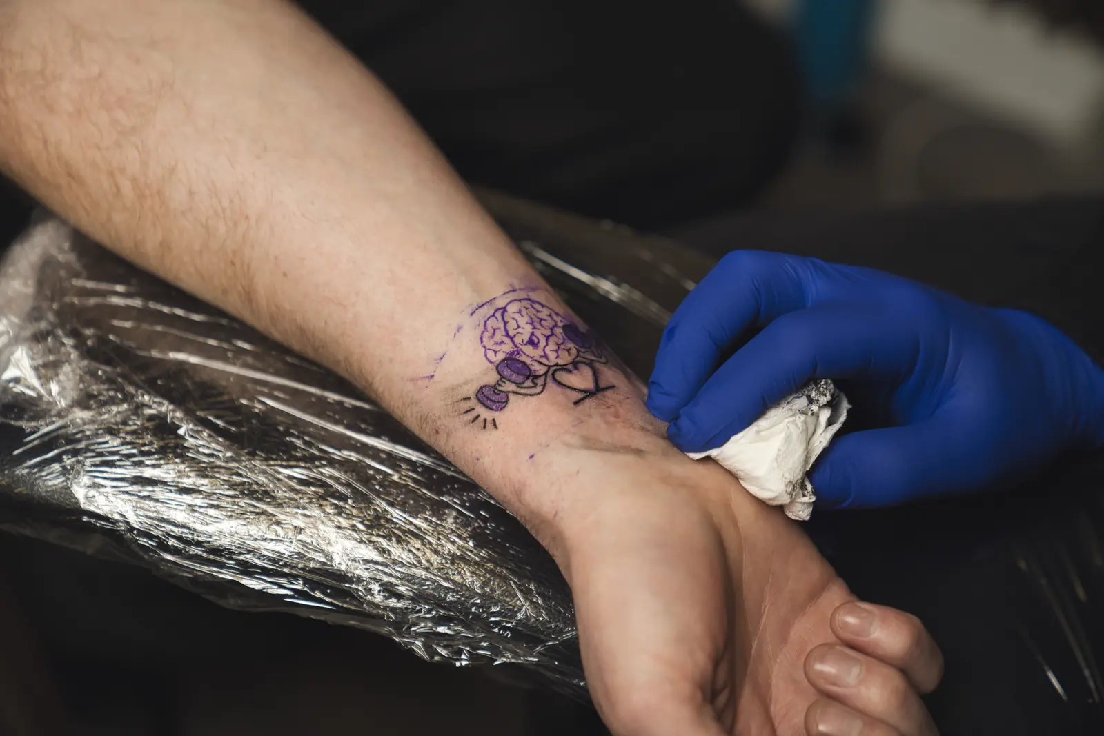A blue-gloved hand wiping a forearm at Gym22. The purple stencil is a cartoon brain lifting dumbbells next to a heart, over clear plastic.