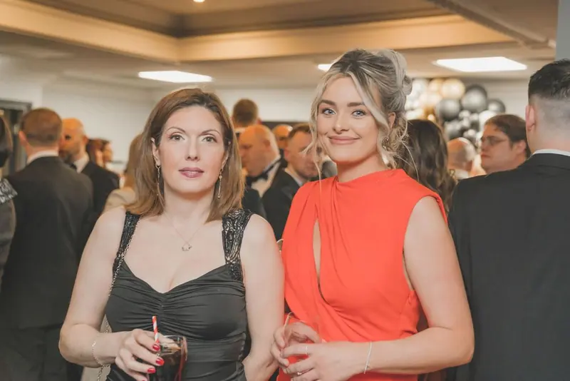 Two women at the Inn Collection Group event in Newcastle, one in a black dress and one in a bright orange dress, with black and gold balloons behind.