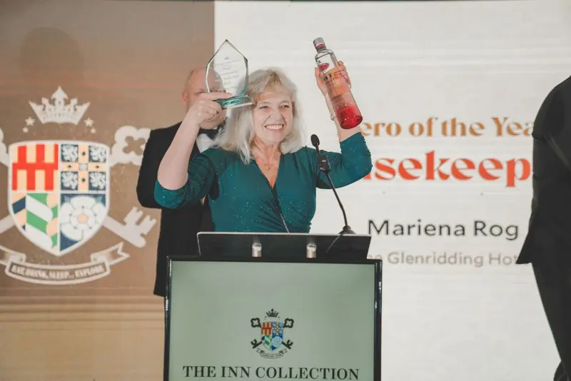 A woman in a sparkly teal dress at a lectern, with a glass trophy and a bottle, and a man in a dark suit behind her. Inn Collection Group, Newcastle.