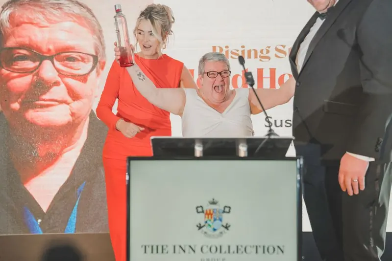 Three people on stage at the Inn Collection Group event in Newcastle: a woman in a bright orange dress raising a bottle, a woman in white, and a man in a black tuxedo.
