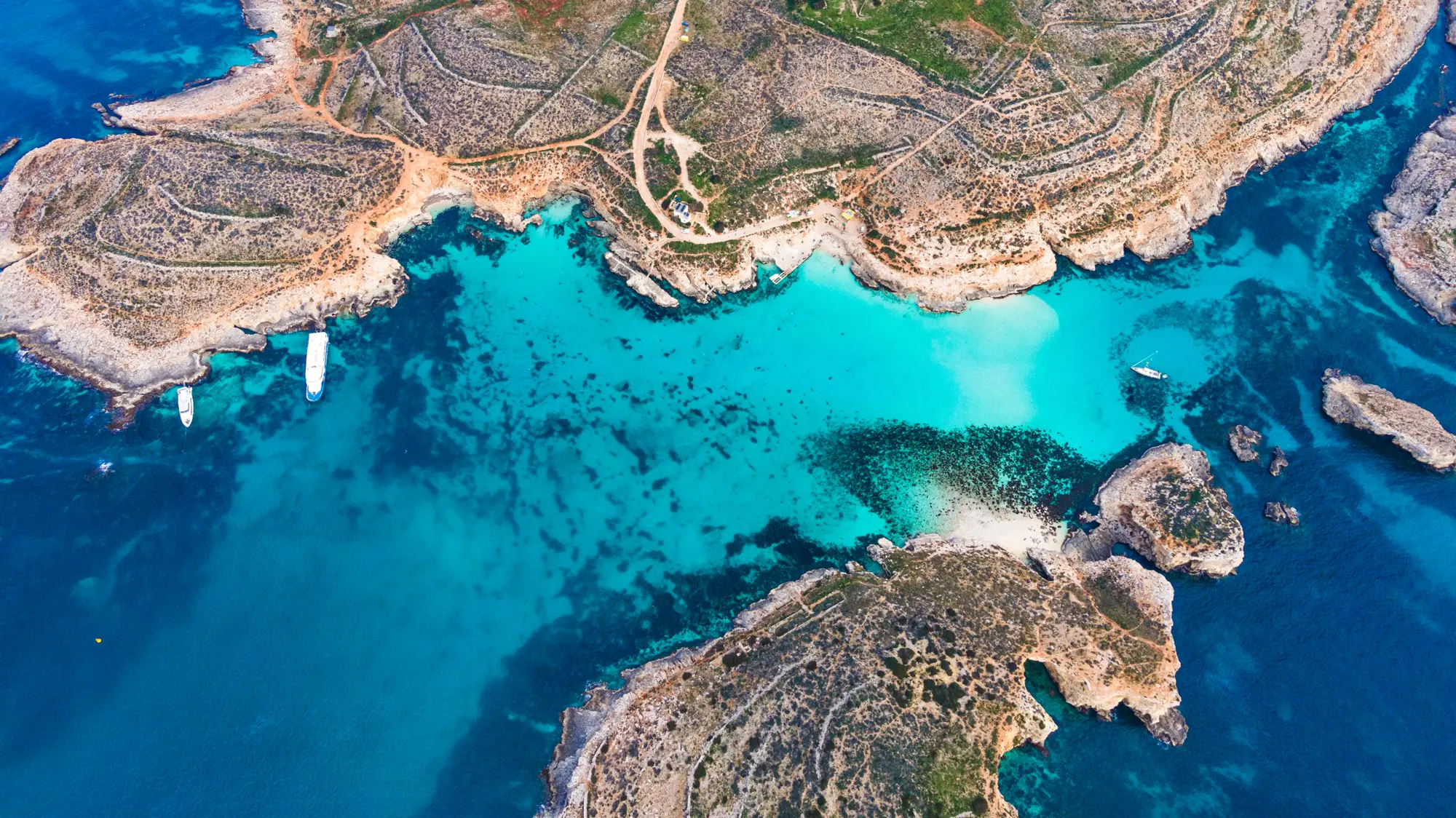 Aerial view of the Blue Lagoon, Comino: rocky ground, dirt tracks, and a bay of turquoise and deep blue water.