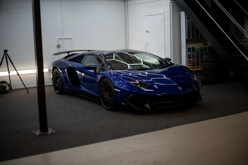A low blue sports car with black wheels, parked indoors on a dark floor against white brick, near a staircase.