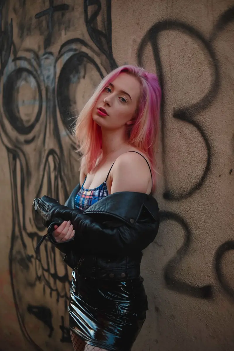 A woman with pink-and-blonde hair, a black leather jacket off the shoulders, a blue plaid top, a black mini skirt, and fishnet tights, leaning on a wall with graffiti.