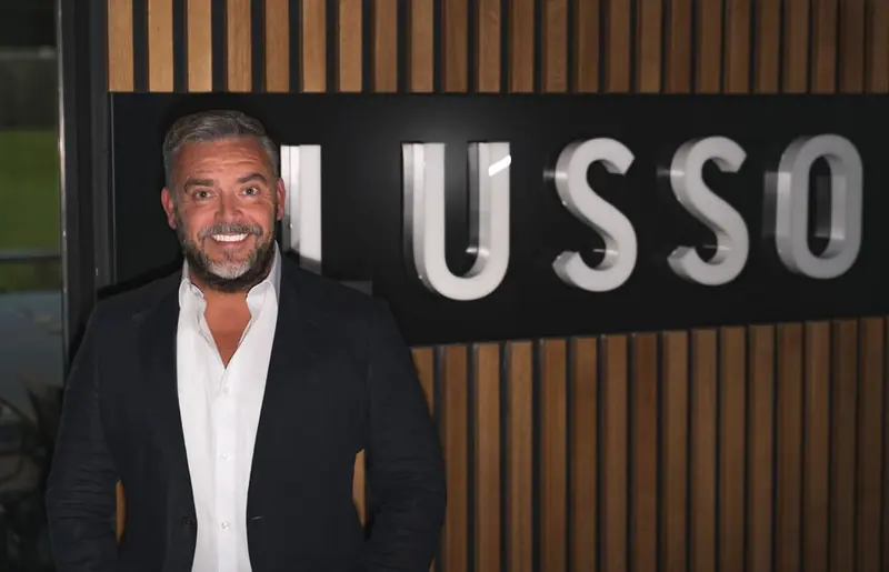 One man at night in a black blazer and an open white shirt, hands in his pockets, in front of a wood-slat wall and an illuminated sign. Lusso Stone.