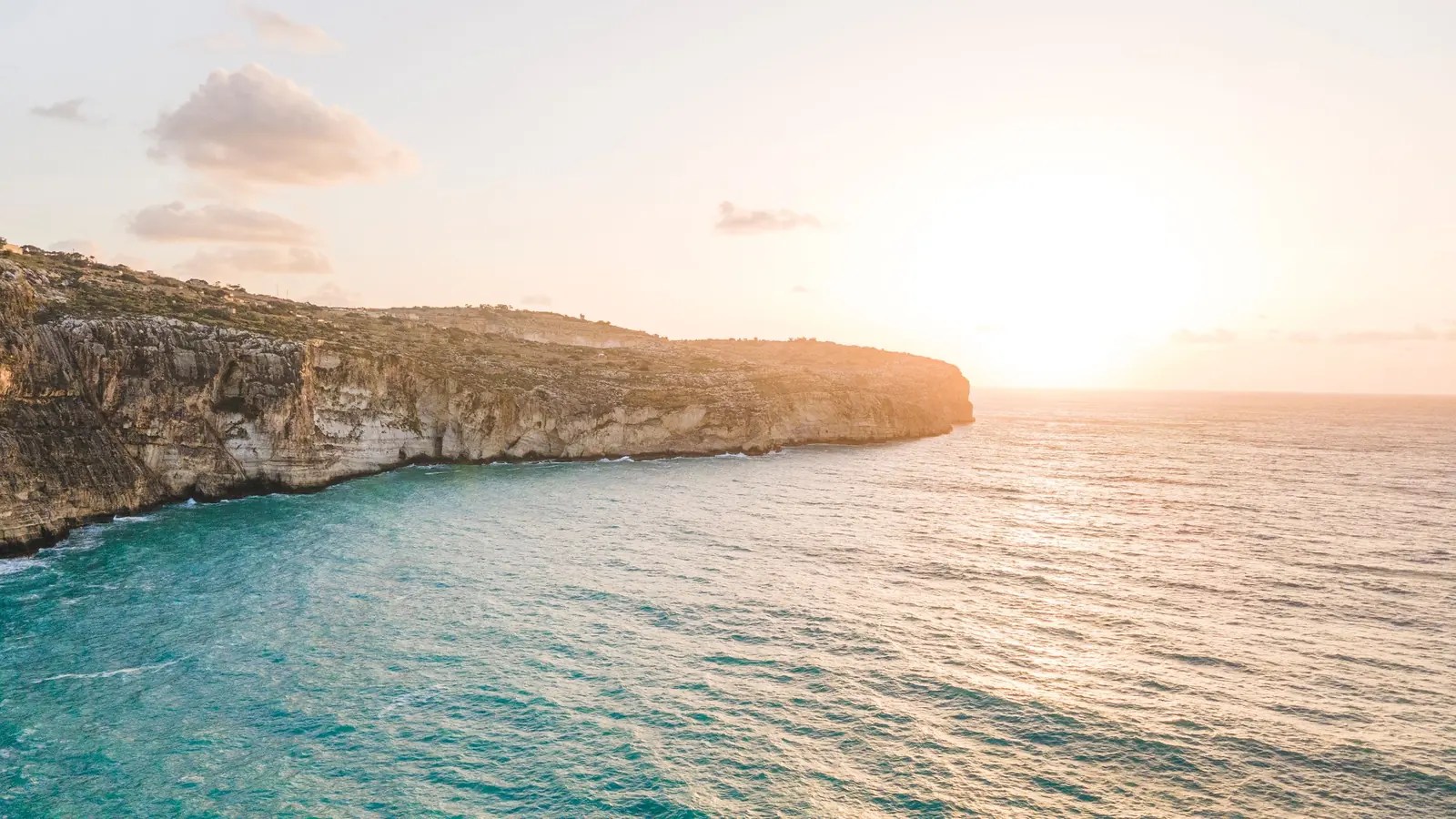 A long pale rocky cliff with scrub beside clear turquoise water, the sea turning deeper blue, in low sun. Malta.