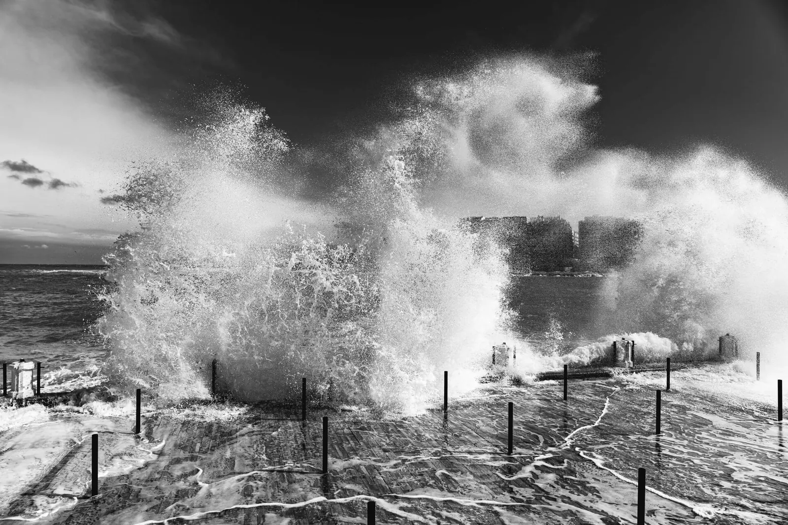 Black and white photograph of large waves breaking over a wet promenade with short posts, spray in the air, and buildings beyond. Malta.