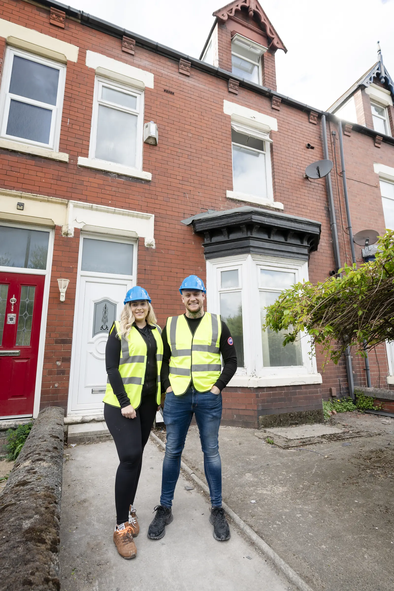 Two people in blue hard hats and yellow hi-vis on a concrete path in front of a red-brick house. MEL Property.