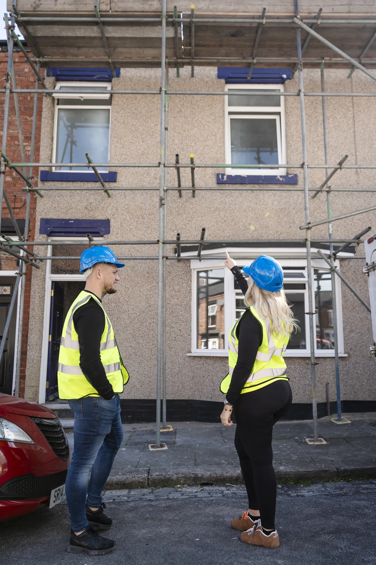 Two people in blue hard hats and yellow hi-vis on a pavement in front of a pebbledash house in scaffolding. One points up. MEL Property.