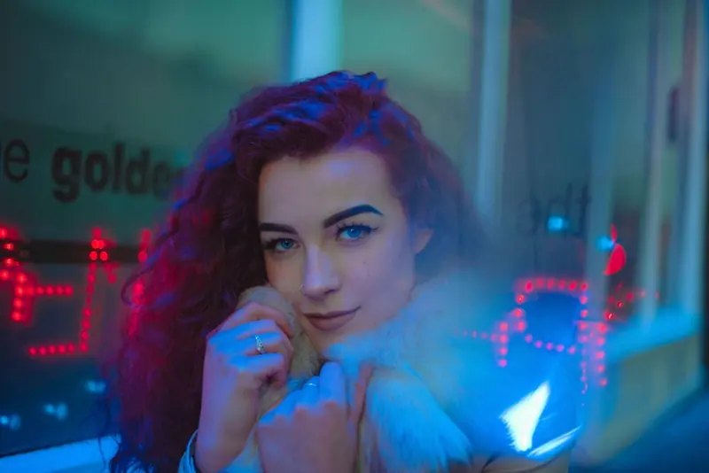 A woman with long curly hair holding a small light-coloured dog to her face, in a coat with a thick white fur collar, outside a shopfront lit blue with rows of red lights.