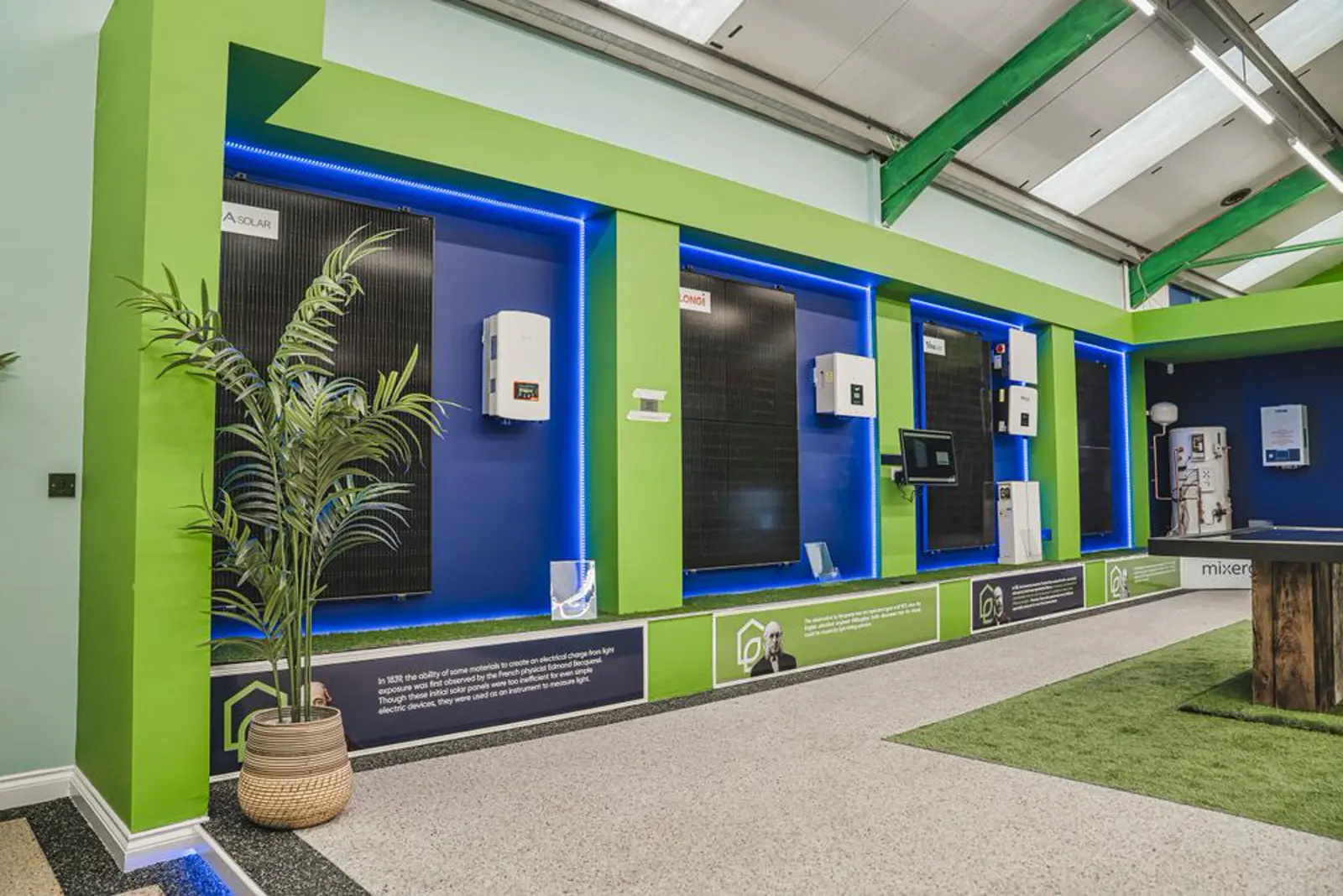 An empty indoor showroom with green pillars, blue display recesses, black solar panels, and white electrical units. Northern Renewable Centre.