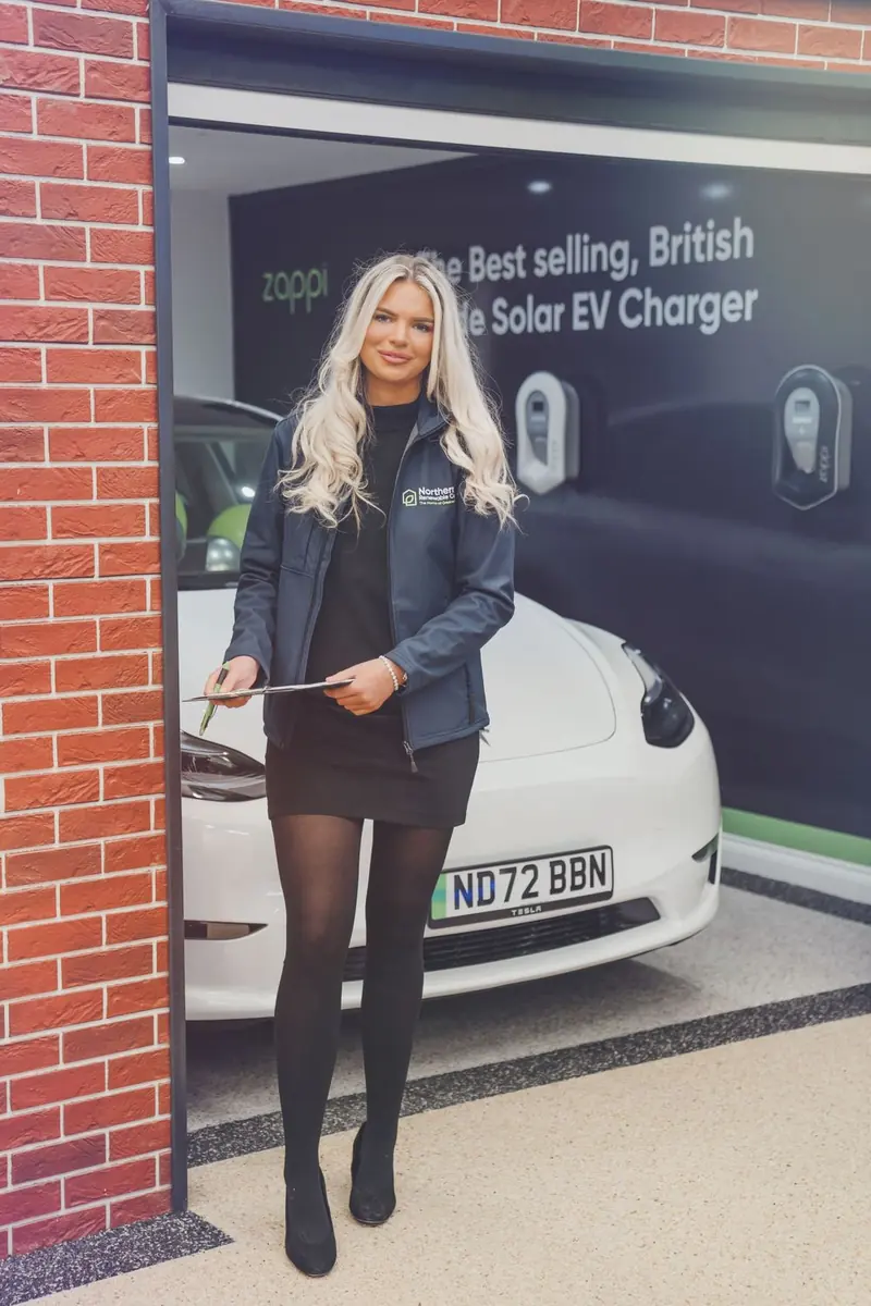 A woman with long blonde hair in a dark blue jacket and black dress, holding a clipboard and pen in a garage doorway, a white car and wall chargers behind her. Northern Renewable Centre.