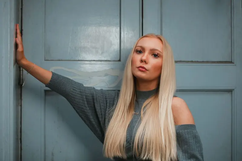 A woman with long blonde hair at a pale blue panelled door, in a grey off-the-shoulder knit, one hand on the frame.