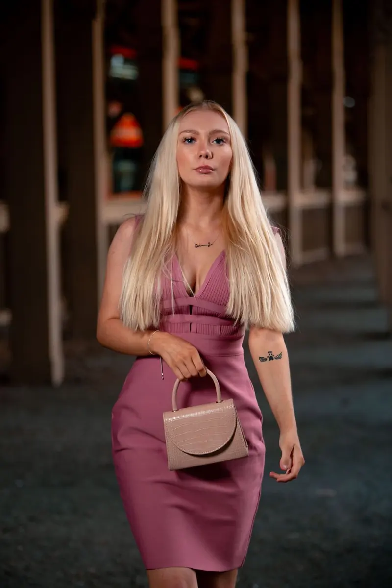 A woman with long straight blonde hair in a mauve sleeveless dress, carrying a beige handbag, walking toward the camera on a dark street at night.