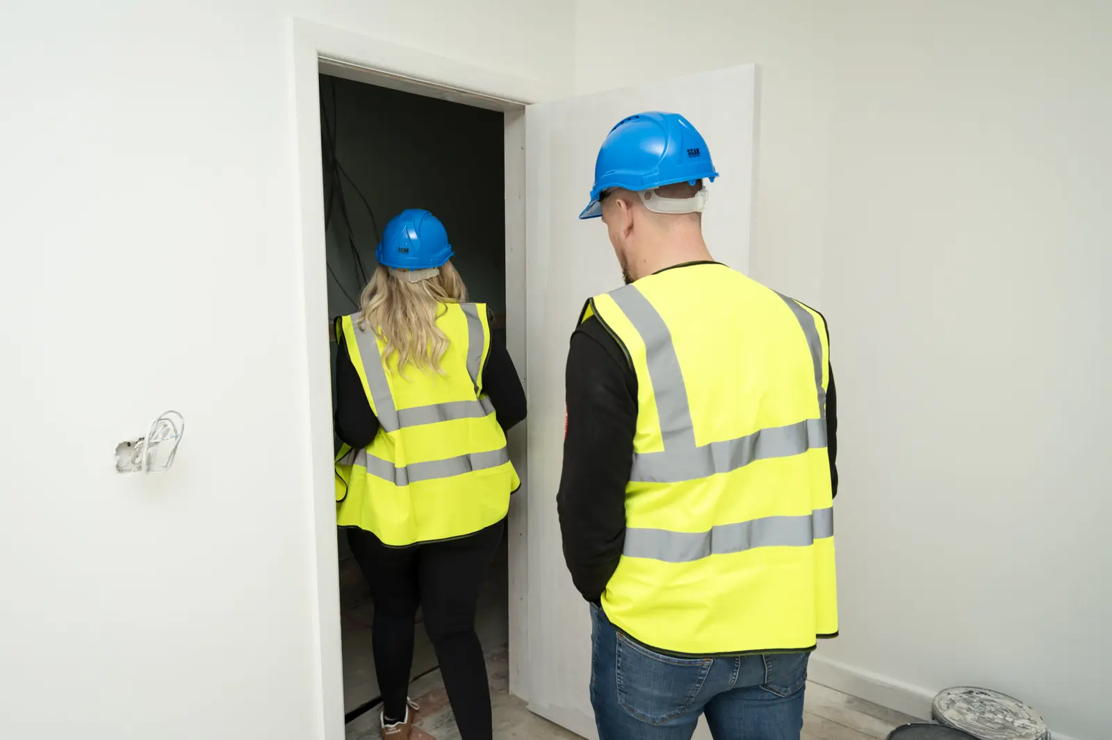 Two MEL Property staff in yellow hi-vis and blue hard hats, seen from behind at an open doorway into an unfinished room.