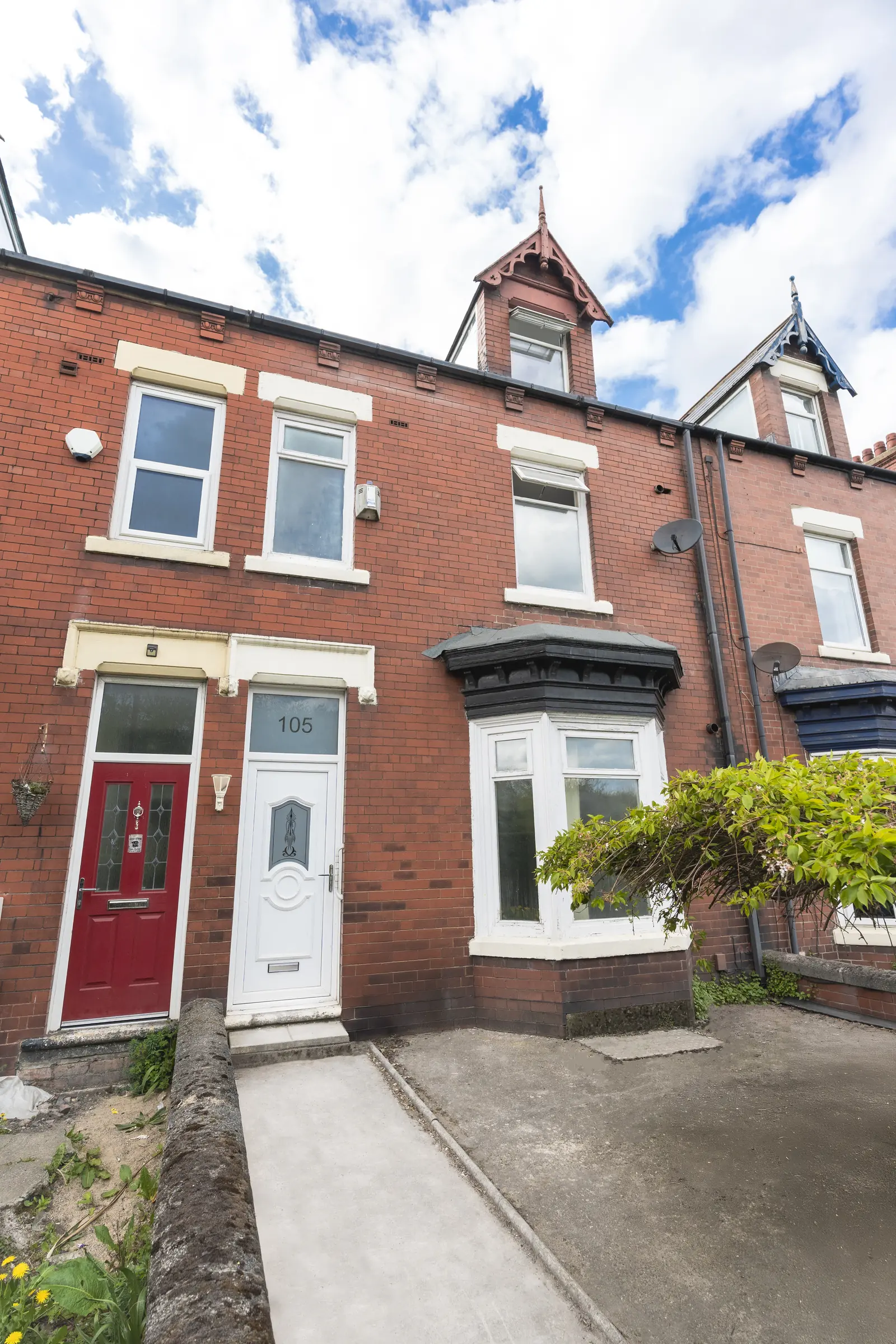 A row of red-brick terraced houses under a blue sky, with a red door, a white door marked 105, and pointed dormers. No people.