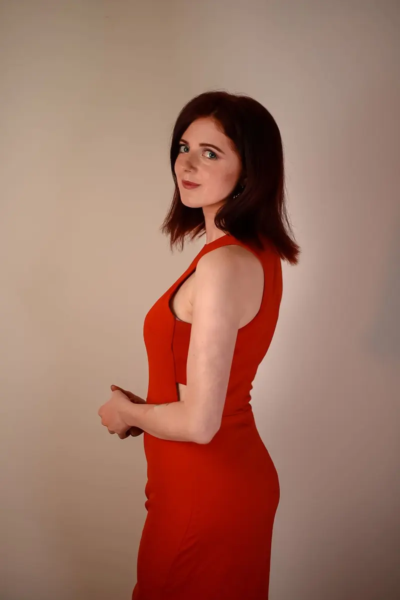 A woman with dark reddish-brown hair in a sleeveless orange-red dress with side cutouts, against a plain beige wall, looking back at the camera.