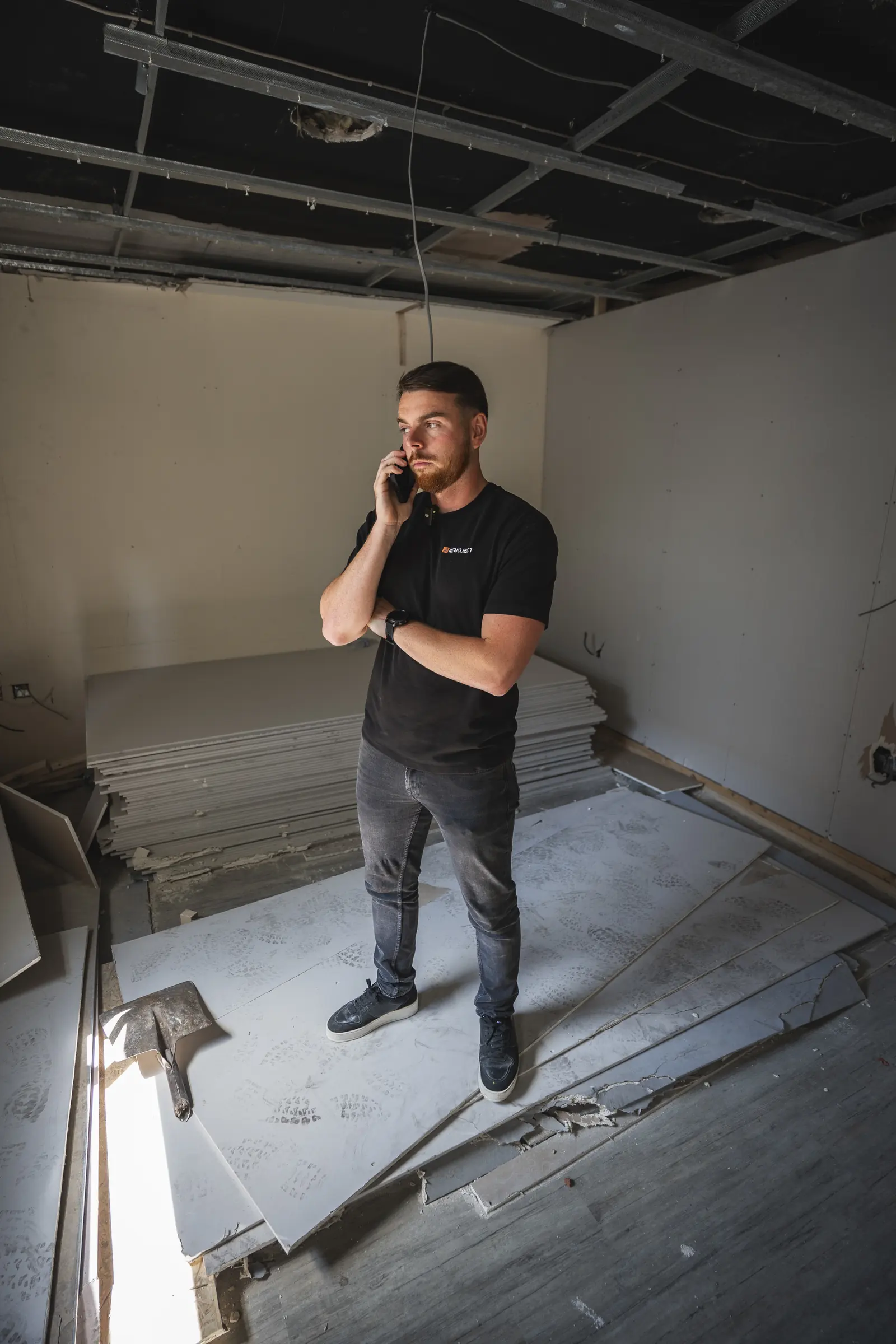 A bearded man in a black T-shirt on his phone in an unfinished room, with stacked plasterboard and an exposed ceiling. Renoject.