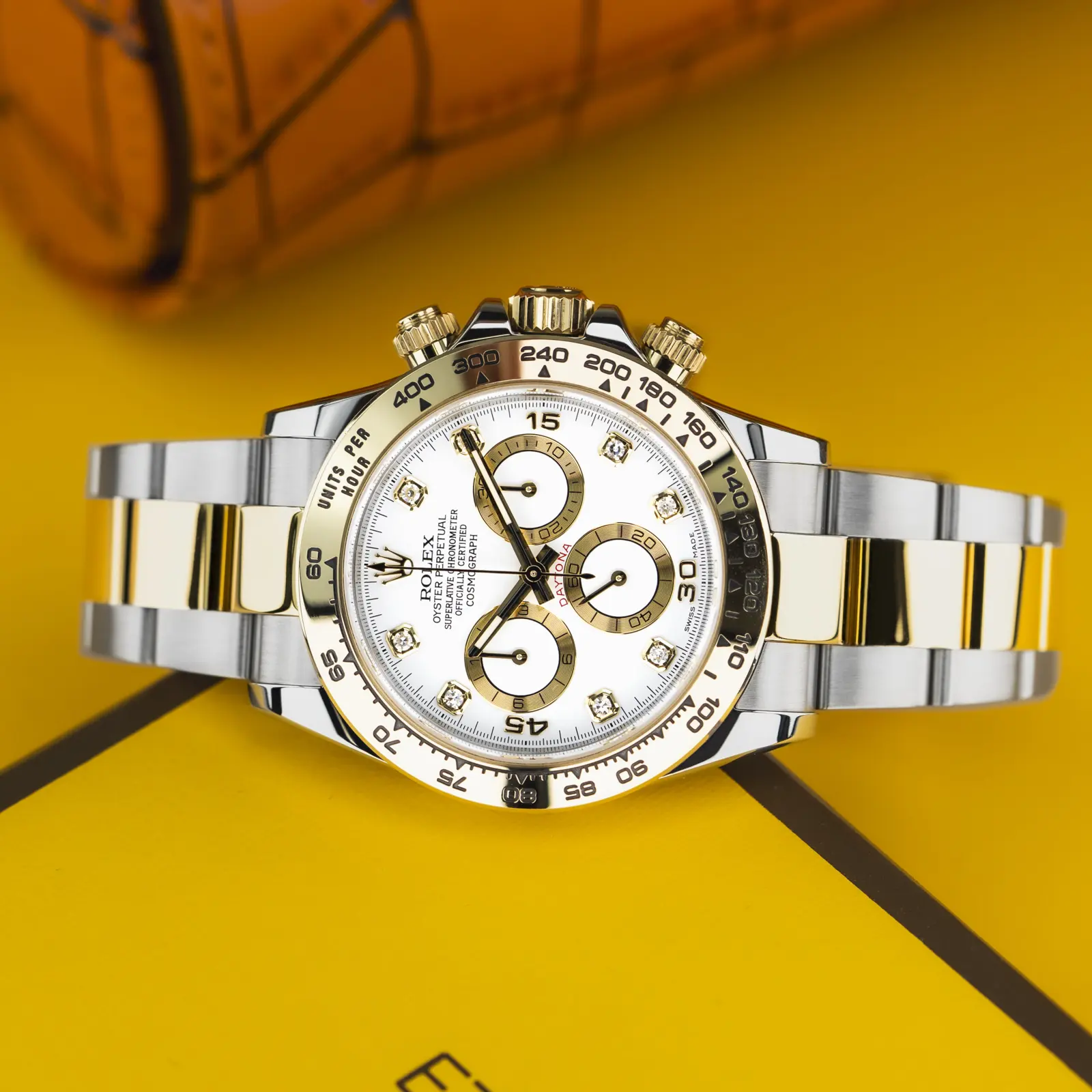 A two-tone silver-and-gold chronograph with a white dial, on a bright yellow surface, with an orange blur behind it.