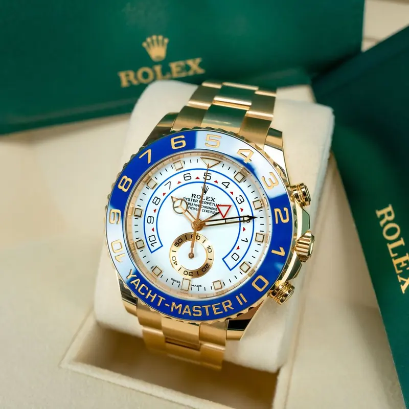 A gold wristwatch with a white dial and a blue bezel, on a cream cushion, with a dark green box behind it.