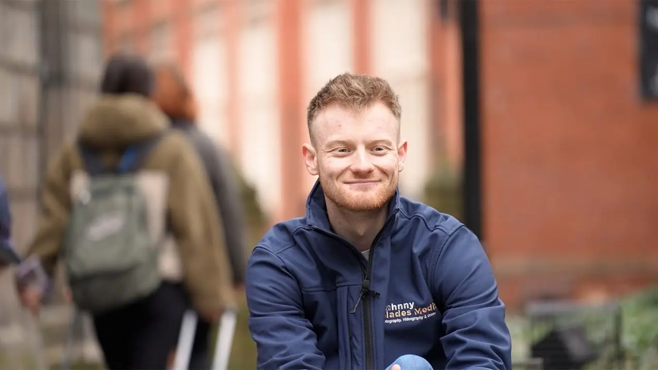 A man in a navy zip jacket outdoors in front of a red brick building, with two blurred figures behind him.