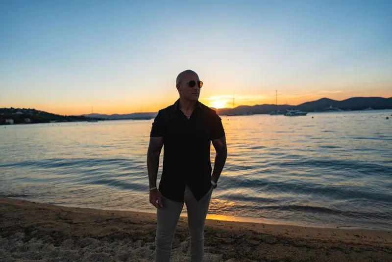 A man on a sandy beach at sunset in a black short-sleeve shirt and light trousers, with water and boats behind him. St Tropez.