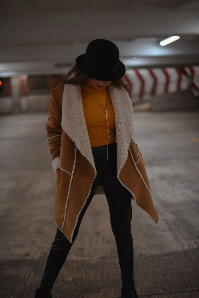 A woman in a car park looking down, in a black bowler hat, a mustard-yellow top, a camel coat with cream shearling, and black leather trousers.