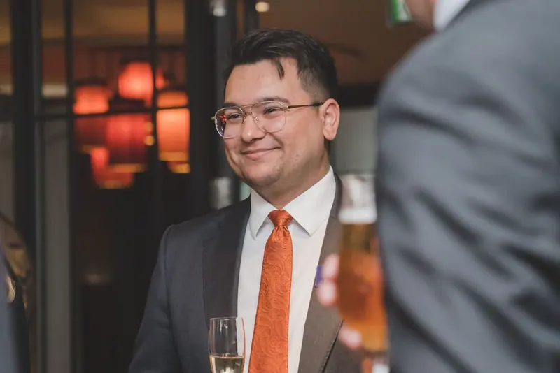 A man in a dark suit, white shirt, and orange tie, holding a pale drink, with another person in a grey jacket seen from behind. Warm red lights.
