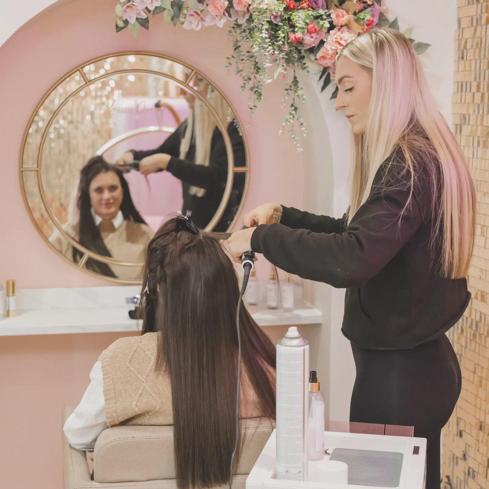 In a pink-walled salon, a stylist with long blonde-to-pink hair uses a heated tool on a seated client with very long dark hair. Kayleigh Jane.
