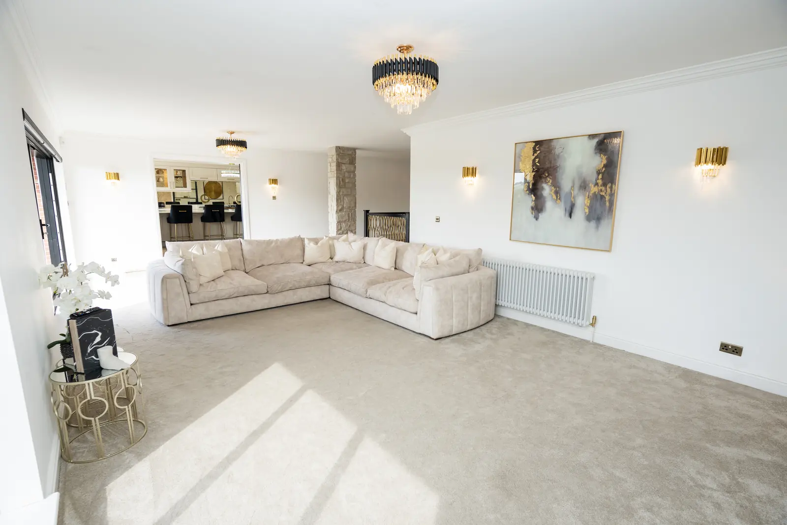 An empty living room at The Green, Thornaby: a cream L-shaped sofa, gold wall lights, a gold-framed painting, and a chandelier, with a kitchen beyond.