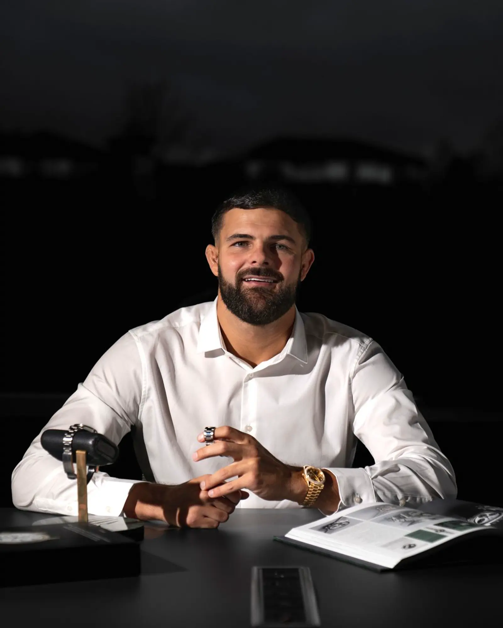 A man in a white shirt at a dark table, a gold watch on his wrist, a second watch on a stand, and an open catalogue. Time Trader.