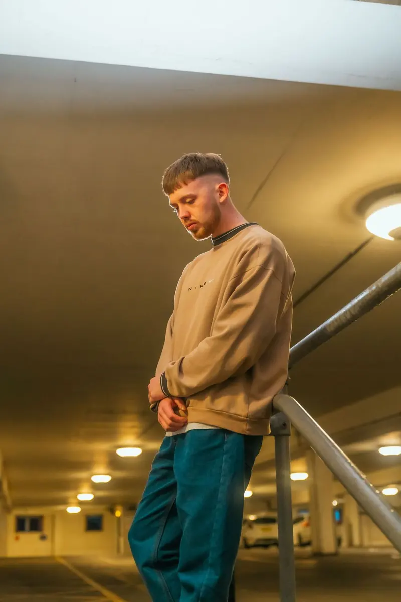 A man with short brown hair and a beard, in a beige sweatshirt and teal trousers, looking down beside a railing in an indoor car park.