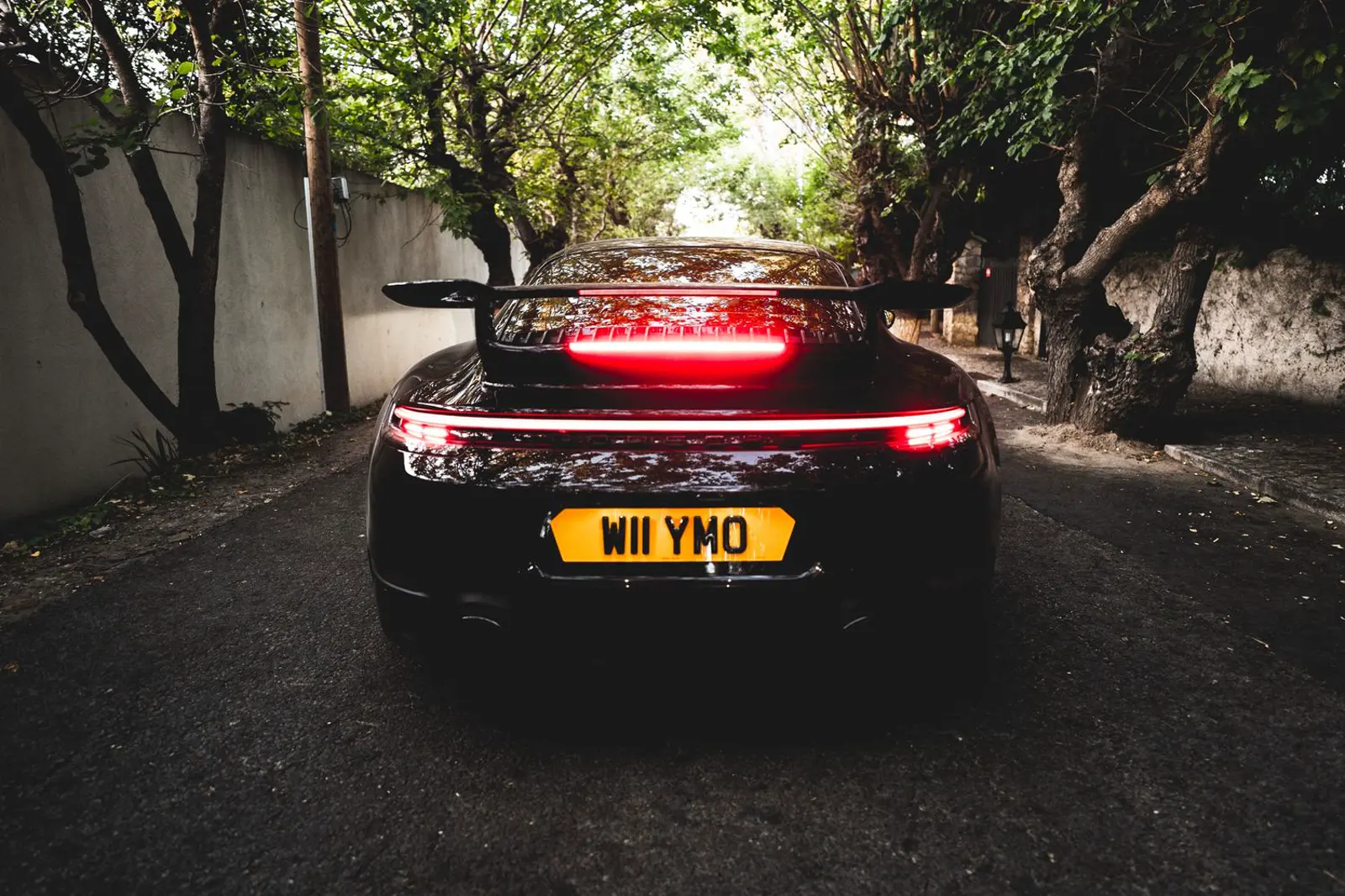 The rear of a black car on a tree-lined street, with a full-width red light bar lit. David Whyman.